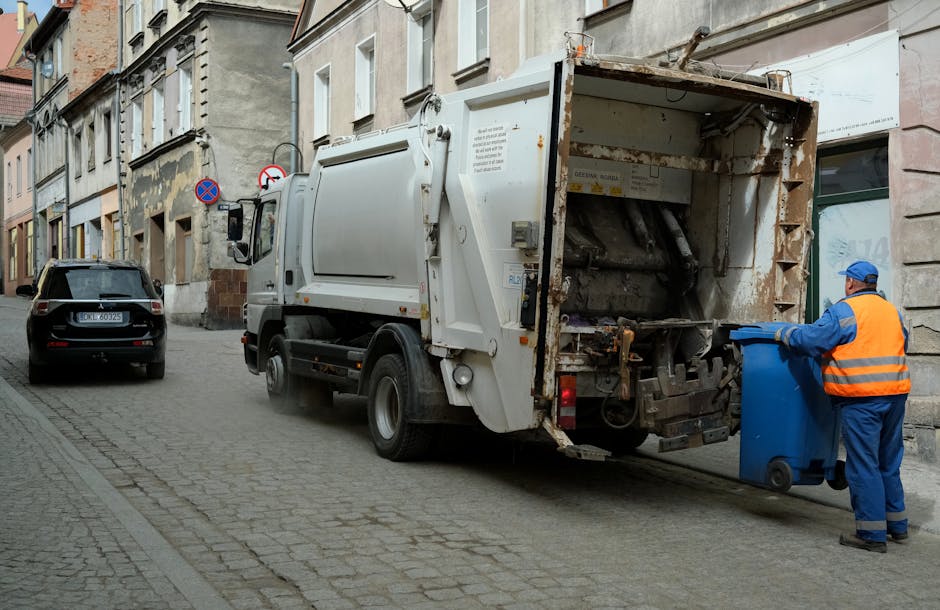 A waste collection vehicle operated by Waste Removal Kennington is parked along a narrow cobblestone street in an urban residential area, with multi-story buildings lining the roadside. The truck has a white, utilitarian body with visible rust and dirt around the loading area, which is open and exposed at the rear. A worker wearing a blue uniform, orange high-visibility vest, and protective gloves is positioned on the right side of the image, actively placing a blue wheelie bin into the truck’s intake compartment. The street features parked cars on the left, including a dark grey vehicle with a visible license plate, and the background showcases aged building facades with a mix of stone and plaster finishes, some showing signs of weathering. Two traffic signs are mounted on the building, indicating parking restrictions and no stopping zones. The overall scene depicts a routine rubbish removal operation suitable for an independent waste disposal service in an urban setting, emphasizing professional, on-site waste collection amidst city surroundings.