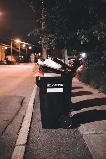 A large outdoor area shows multiple overflowing refuse bins and scattered rubbish bags on a paved surface, with adjacent parked cars and a metal railing. The collection includes black, red, and green bins, some open and filled with mixed waste, alongside various discarded cardboard boxes, plastic bags, and packaging materials. Behind the bins, there is a building with a storefront that has a sign for a fish bar and other retail outlets, partially obscured by a storefront awning and blue construction or scaffolding on the upper level. The waste, consisting of papers, cardboard, plastic containers, and general household waste, appears to have accumulated beyond the capacity of the bins, spilling onto the surrounding pavement. The environment suggests an urban or commercial area, where waste removal may be required to prevent littering or collection delays, reflecting the importance of professional rubbish services such as those provided by Waste Removal Kennington for effective on-site clearance and waste management.