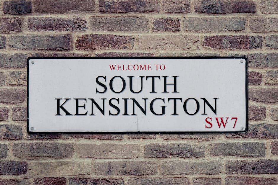A rectangular metal street sign mounted on a red brick wall, displaying the text 'Welcome to South Kensington SW7' in black and red lettering. The sign features a white background with black borders, and the brick wall behind it consists of uniformly shaped bricks with a rough texture and varying shades of red and brown. The sign is positioned centrally on the wall, with visible screw fixings at each corner. The setting suggests an outdoor location, likely on a building facade in a residential or commercial area of South Kensington, with natural daylight illuminating the scene. This type of signage is often associated with area identification or private property markers, relevant in contexts of urban waste management and alternative disposal options, such as private rubbish or bulky waste collection services provided by Waste Removal Kennington.