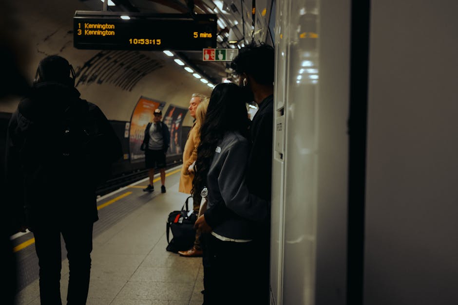 In the foreground, a person with dark curly hair wearing a dark jacket is standing next to a pillar in a subway station, partially obscuring their face. To the left, another individual with a backpack, dressed in dark clothing, is walking away from the camera along the platform. In the background, several passengers are standing on the platform, some with luggage or bags, waiting for their train. The platform has a smooth, grey surface with yellow safety markings along the edge. Overhead, a digital train schedule display shows upcoming train destinations, including Kennington, with the time remaining until arrival, illuminated against the tunnel's curved ceiling. The station is lit with bright, fluorescent lighting reflected on the polished surfaces of the pillars and the station’s walls. The environment appears clean, modern, and organized, typical of an urban underground transit system. This scene captures the typical activity at a busy underground station, relevant to discussions about urban transportation and private or alternative transportation options, which can be linked to the broader context of waste management infrastructure and service accessibility by Waste Removal Kennington for related rubbish collection needs.
