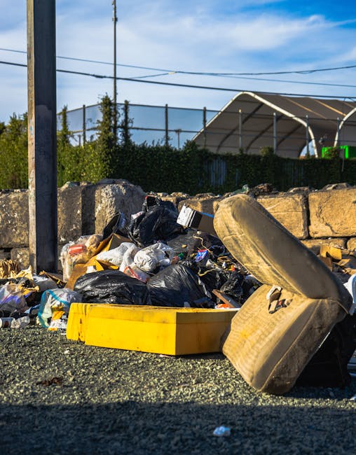 An outdoor scene showing a collection of mixed waste and rubbish accumulated on a gravel surface, including black plastic bin bags filled with various items, an old, worn car tire lying on its side, and a yellow plastic container or tray partially obscured by the debris. The rubbish appears to include paper, plastic packaging, and miscellaneous refuse, indicating unsorted waste awaiting collection or disposal. In the background, there's a stone wall constructed from uneven, weathered stones, with some greenery such as bushes or small trees behind it. To the left, a metal pole supports overhead power lines, and further back, a sports or tennis court with a high fence and a curved, semi-transparent roof structure is visible under a bright blue sky with scattered clouds. The scene suggests an environment where private rubbish collection services, like those provided by Waste Removal Kennington, may handle on-site waste clearance outside conventional municipal waste systems.