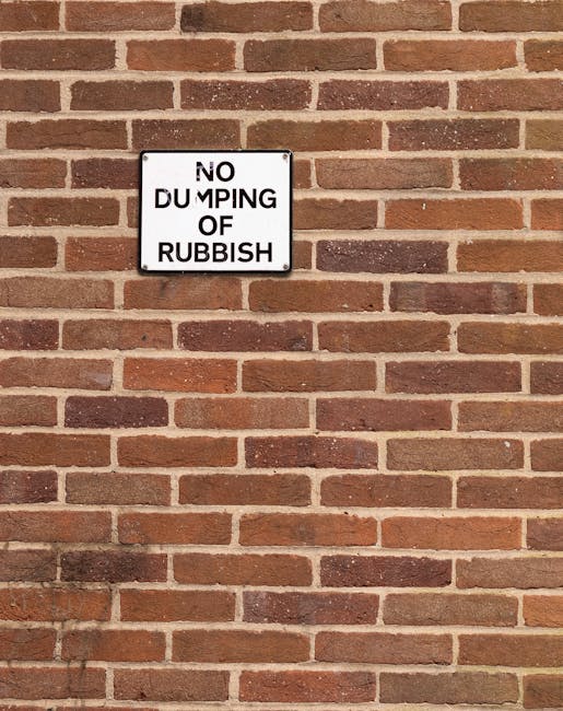 A rectangular black and white sign mounted on a red brick wall with a textured surface. The sign reads 'NO DUMPING OF RUBBISH' in bold black lettering, with the word 'DUMPING' partially obscured due to damage or wear. The brick wall behind the sign consists of evenly laid bricks in varying shades of reddish-brown, with light gray mortar joints. The overall scene appears to be outdoors, with the sign serving as a notice to prevent illegal rubbish dumping in the area. The clarity of the sign indicates it may be part of a local waste management effort or private property boundary to discourage rubbish disposal and promote proper rubbish removal practices, relevant to services provided by Waste Removal Kennington.