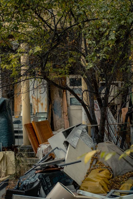 A mixture of discarded household items and debris is seen piled on the ground outdoors, with various materials including wooden panels, an old white appliance, and metallic objects. The pile appears disorganized and is situated near a tree with dark, twisting branches and green foliage, partially obscuring a background that includes weathered building walls with peeling paint and rusted metal surfaces. Some items are covered with grey fabric or plastic sheets, and a yellow bag is visible among the waste materials. The scene is illuminated by natural light, with shadows cast by the surrounding trees and objects, suggesting an outdoor setting on an overcast day. This cluttered area exemplifies the type of waste that waste removal services such as Kennington-based rubbish collection companies might collect during private clearance or on-site rubbish removal, emphasizing the need for professional disposal of bulky household waste.