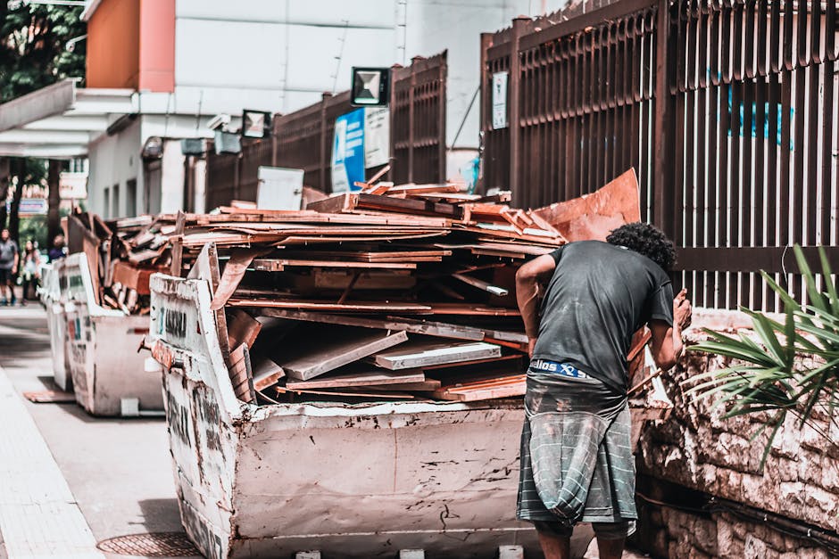 A man with curly black hair, wearing a dark grey T-shirt and a plaid kilt, is standing on a sidewalk next to a large, rusty, white skip filled with wooden planks, panels, and construction debris. The wooden materials vary in size, with some planks leaning outward and others stacked horizontally, displaying a mix of weathered brown and reddish tones with rough textures. The skip is positioned beside a stone wall with a spiky leafy plant growing nearby. In the background, there is a tall brown metal fence, partially obscuring buildings that include signage and a street sign. The scene is outdoors during daylight, with natural light illuminating the area, and a few pedestrians visible further along the sidewalk. This image depicts a typical scene of DIY or construction waste disposal suitable for private rubbish removal services like Waste Removal Kennington, which specializes in efficient on-site clearance for builders and commercial clients.