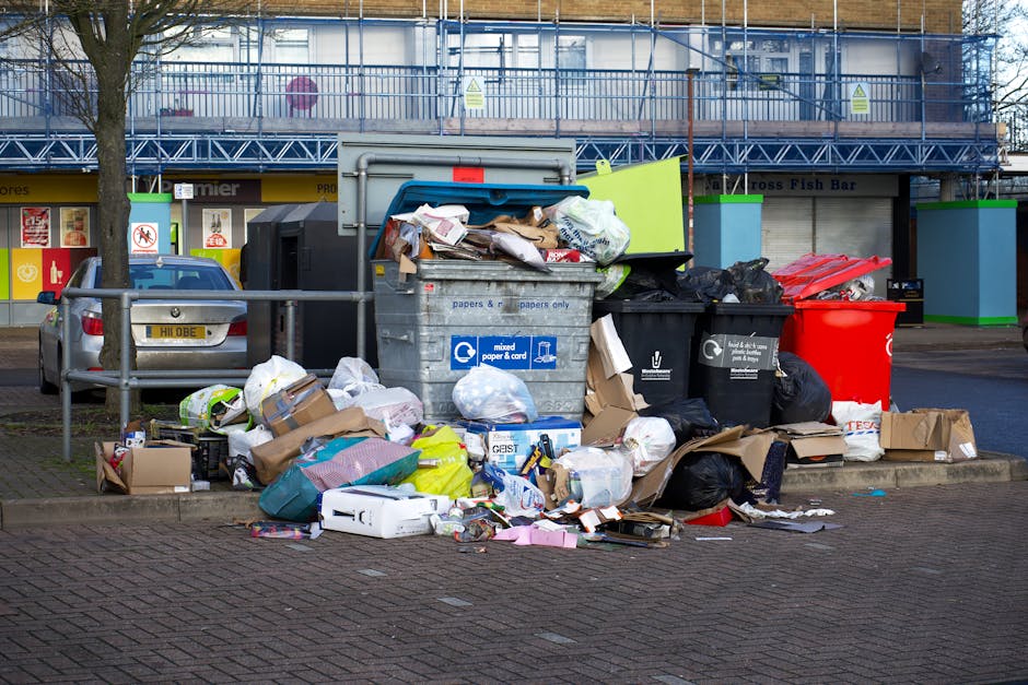 A large collection of mixed waste items and refuse, including cardboard boxes, plastic bags, paper bundles, and various discarded packaging materials, are piled on a paved area beside a parking space. The waste is overflowing from multiple black and black-and-white recycling bins, with some containers leaning and spilling onto the ground. Behind the waste, there are two parked cars, one silver and one dark-colored, with the silver car partially obscured by a metal railing. The scene is set in an urban area, adjacent to a building with a yellow storefront on the ground level, and a blue scaffolding structure covering the upper part of the building, indicating ongoing construction or maintenance. The overall environment appears cluttered due to an accumulation of refuse, typical of private waste collection sites where rubbish is sometimes temporarily stored before disposal or recycling, with the details emphasizing the need for professional rubbish removal services like those provided by Waste Removal Kennington. The lighting is natural, and the setting suggests daytime, with clearly visible textures of the various materials, reflecting a typical urban rubbish disposal scene.
