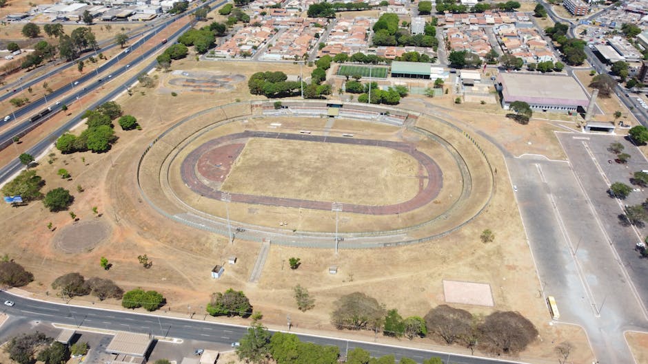 An aerial view of a large, open sports field with a central grass area surrounded by a red running track. The field is situated in an urban area with a mixture of residential and commercial buildings visible in the background. To the left of the field, there are trees and open grassy spaces, some with small patches of dirt. A paved pathway runs along the perimeter of the sports track, with a few small structures and light posts positioned around the field. Adjacent to the sports area, there is a parking lot with a yellow vehicle parked, and the surrounding streets show minimal traffic. The environment is well-lit, indicating daytime with clear weather, and the scene is calm and undisturbed, with no visible activity on the field. This setting illustrates a community sports facility that may be accessible for outdoor activities, and the empty field and track might be suitable for private or alternative sports and recreation use, aligning with local waste management and clearance services that handle site cleanups or sports facility maintenance.