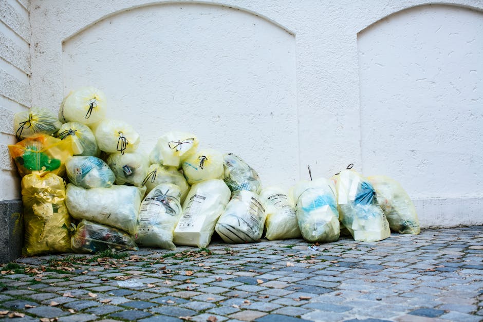 A collection of tightly packed black, yellow, and clear plastic rubbish bags filled with waste, stacked against a white exterior wall. The bags appear to contain a mixture of household refuse, some with visible labels or packaging, and are secured with black ties or knots. They are placed on a cobblestone pavement outdoors, with some small scattered debris and leaves at the base of the pile. The background shows the textured, painted surface of a plain white wall, with the bags leaning slightly to the left side of the image. The scene suggests an area designated for rubbish collection or private waste disposal, commonly handled by rubbish removal services like Waste Removal Kennington, providing an alternative to official council bins. The natural daylight highlights the different textures of the plastic bags and the uneven surface of the cobblestones, emphasizing the practicality of on-site waste management.