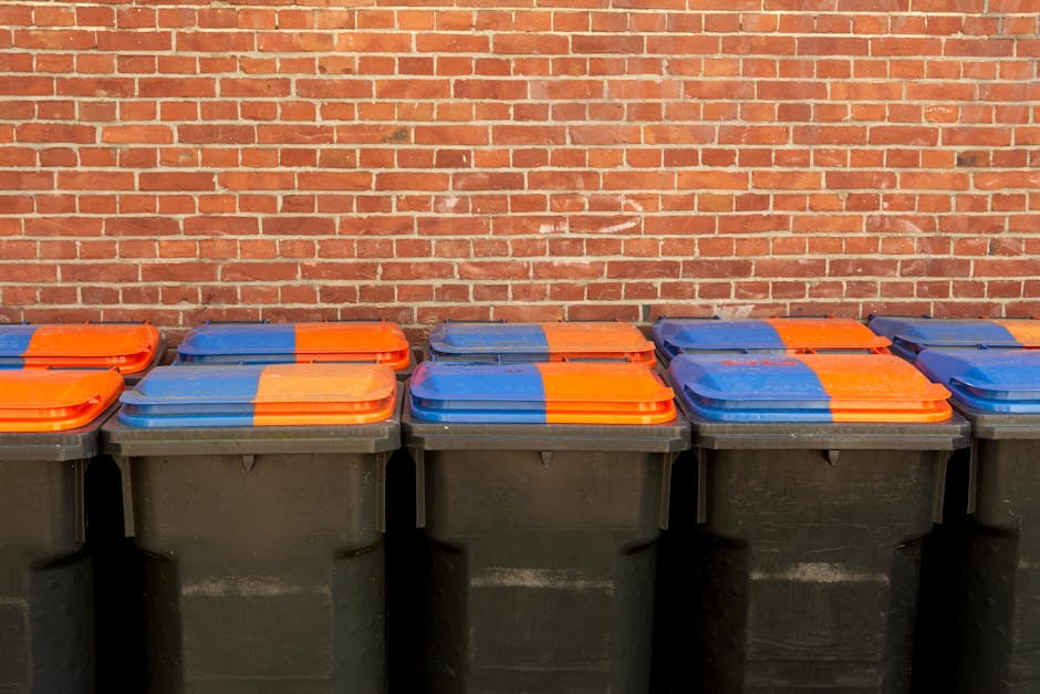 A collection of tightly packed black, yellow, and clear plastic rubbish bags filled with waste, stacked against a white exterior wall. The bags appear to contain a mixture of household refuse, some with visible labels or packaging, and are secured with black ties or knots. They are placed on a cobblestone pavement outdoors, with some small scattered debris and leaves at the base of the pile. The background shows the textured, painted surface of a plain white wall, with the bags leaning slightly to the left side of the image. The scene suggests an area designated for rubbish collection or private waste disposal, commonly handled by rubbish removal services like Waste Removal Kennington, providing an alternative to official council bins. The natural daylight highlights the different textures of the plastic bags and the uneven surface of the cobblestones, emphasizing the practicality of on-site waste management.