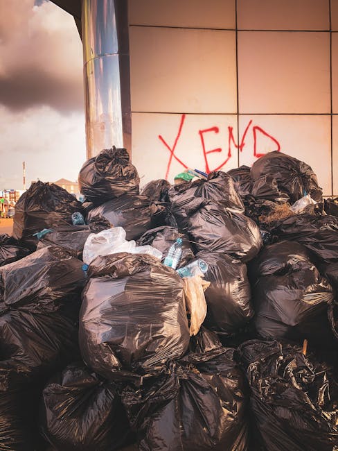 A large pile of black rubbish bags, some with visible knots and creases, stacked against a modern tiled wall in an outdoor urban environment. Several clear plastic water bottles are protruding from the bags, indicating typical household waste. Behind the bags, the wall features a prominent red graffiti spray-painted with the partially visible word 'XEND'. To the left, part of a metallic cylindrical structure reflects the surrounding light, while a cloudy sky with patches of daylight is visible above the scene. The area appears to be a designated waste disposal site or informal rubbish collection point, illustrating the challenges of managing waste outside of scheduled council collection, as handled by waste removal services such as Waste Removal Kennington.