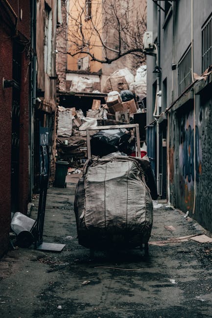 A narrow alleyway in an urban area cluttered with piles of mixed waste and rubbish bags, with a large black and grey trash bag prominently placed in the foreground. The alley is bordered by tall buildings on both sides, with graffiti visible on the right wall and a small vent or electrical panel affixed to it. In the background, there are overflowing cardboard boxes and miscellaneous discarded items stacked against a wall, indicating a significant accumulation of waste. The ground appears stained from previous spills or dirt, and a tipped-over bin can be seen on the left side of the alley. Leafless tree branches extend overhead, suggesting the scene is set in late autumn or winter. The environment suggests an area needing on-site rubbish clearance, with Waste Removal Kennington potentially providing an alternative waste handling solution for such restricted access or urban settings, supporting efficient private disposal services.
