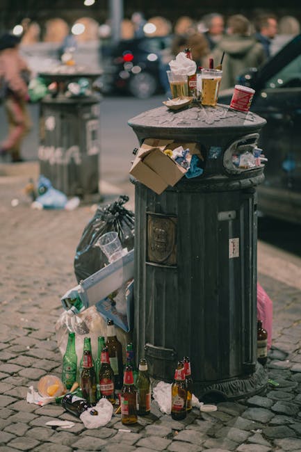 A black metal rubbish bin with a domed lid is situated on a cobbled sidewalk in an urban environment. The lid of the bin is topped with various discarded items, including crumpled paper, empty plastic cups with attached plastic straws, and other small debris. Attached to the side of the bin are cardboard boxes and plastic bags holding additional waste. Surrounding the base of the bin, several glass bottles, some with liquids inside, are strewn on the ground along with crumpled paper and other small litter. In the background, a blurred city street scene is visible with people walking and interacting, along with parked vehicles and a grey trash container. The environment appears to be lively with natural daylight, and the scene reflects urban littering typical of a public gathering area, emphasizing the need for waste removal services like those provided by Waste Removal Kennington, especially for street cleanup and waste management in busy city locations.