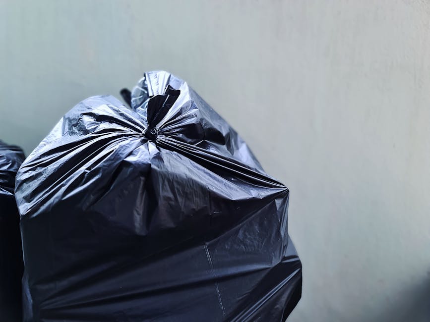 Close-up of a tightly knotted black plastic rubbish bag with a smooth, glossy surface showing subtle creases and folds. The bag is positioned in the foreground against a plain, light-colored wall, with additional black bags partially visible behind it. The bags appear to be filled with waste, possibly household refuse, and are placed on a solid surface, which may be a floor or pavement. The scene suggests an outdoor or indoor setting suitable for waste collection or disposal, emphasizing the importance of proper rubbish management. The neutral lighting highlights the reflective nature of the plastic material without casting harsh shadows, aligning with the context of private waste collection or on-site clearance services offered by Waste Removal Kennington, supporting alternative rubbish removal methods outside of standard local authority collection schedules.